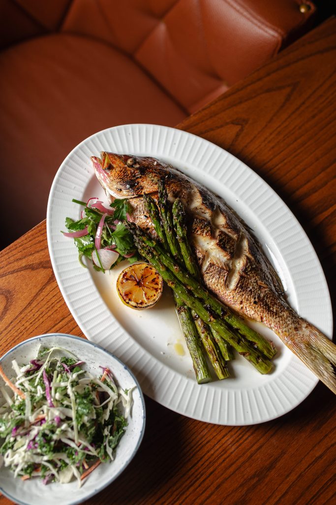 Roasted Whole Fish with lemon and asparagus on a dish, served with a side dish of Tricolor Kale Wasabi Slaw